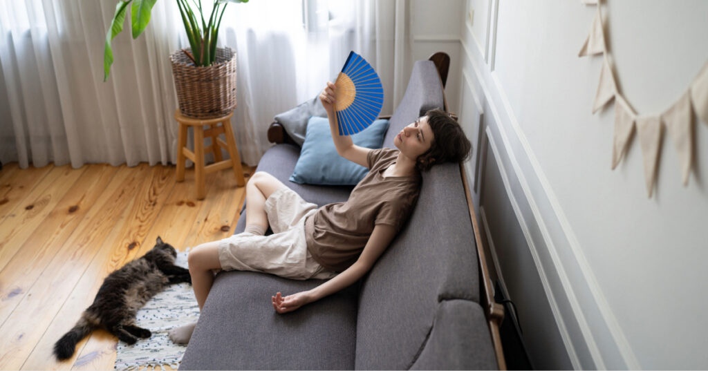 female owner fanning herself on the couch while cat lays at her feet to prevent cat heatstroke