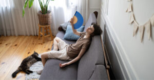 female owner fanning herself on the couch while cat lays at her feet to prevent cat heatstroke